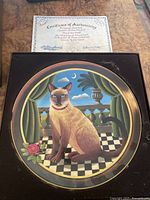 Plate showing a brown cat sitting on checkered floor with a rose and balustrade background, accompanied by certificate of authenticity