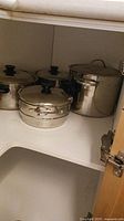 Photo showing stainless steel pots with glass lids arranged inside corner cabinet including stock pot, sauce pan and smaller pots