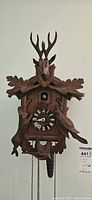 Front view of wooden cuckoo clock with carved deer head, antlers, hunting animal figures, pinecone weight, and Roman numeral clock face