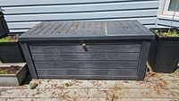 Front view of a large black plastic deck box with horizontal slats, some dirt and leaves on top, lock in front.