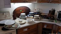 Wide angle photo showing various stacks of plates, bowls, knives in block, and kitchen utensils arranged on white kitchen counters.