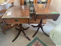 Two matching wooden drop leaf side tables with glass tops and drawers, standing side by side, showing front and side views.