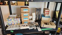 Wide view of shelf containing multiple boxes and packages of vintage intercom interfaces, Halo Power-Tec box, smaller electrical components, and various packages.