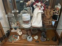 Two glass cloches, ceramic beer stein, three doll figurines, soap stone box and various decorative rocks arranged on two shelves inside display cabinet with mirrored background.