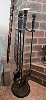 Front view of fireplace tool set showing shovel, broom, and poker on wrought iron stand against brick wall.