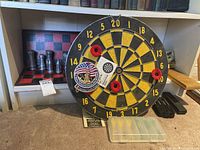 Dart board with 4 darts attached plus 2 more darts in plastic case and metal chess set visible behind the board.