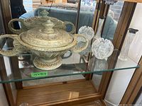Indian decorative bowl with two handles and lid, ornate etched pewter or brass metallic finish, placed on glass shelf.