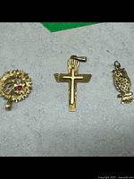 Three gold charms including a floral charm with red stone, a cross with bird design, and an owl charm.