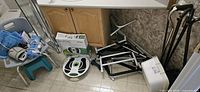 Photo showing multiple mobility aids including Revitive circulation booster, disassembled walker, and white plastic wash basin on tiled floor next to wooden cabinetry.