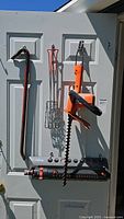 Three garden tools hanging on a door: crow bar, metal sprinkler, and Black and Decker hedge trimmer