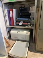Shelf showing printer underneath, binders, toner cartridges stacked above, and loose plastic binder sheets in front