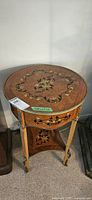 Front angled view of round wooden inlaid table showing floral design on top, brass rim, three legs with metal accents, and lower shelf with matching inlay.