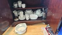 White Johnson Brothers everyday china set arranged in wooden cabinet with dinner plates, side plates, mugs, bowls and casserole server on shelf and cutlery in glass
