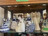 Shelf with assortment of porcelain figurines including KPM and Jim Shore characters displayed against mirrored background.
