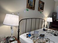 Photo showing one of the table lamps on a side table next to a bed with various jewelry items nearby. Lamp has a shiny metal base with glass section and a white fabric shade. Lamp is lit.