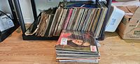 Photo showing a large collection of vinyl records stacked on a floor rack with some additional records in a stacked pile in front, covers are mostly visible and show a mixture of artists and designs.