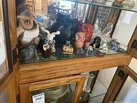 Collection of owl figurines displayed on glass shelf with mirrored background, includes larger ceramic owl and multiple smaller owl figures in varied materials and colors