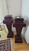 Two dark reddish-brown wooden display pedestals with trapezoidal tapered shape and carved decorative panels, standing on hardwood floor near rug and window.
