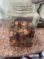 View of clear square plastic container half filled with pennies on a marble-like surface.