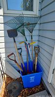 Garden tools including two green metal fan rakes, one green plastic fan rake, shovels, and other garden hand tools in blue plastic bin (bin not included).