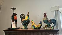 Photo showing wood frog figurine on stand, two wood roosters, ceramic chicken, and ceramic rooster all arranged in a row.