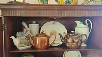 Full shelf view showing the Sadler Old Mill teapot, brown pottery teapot, Wedgwood pitcher, QEII plate and cream teapot arranged on wood shelf.