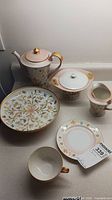 Photo of entire china set arranged including teapot, sugar bowl, milk creamer, cake/sandwich plate, and cup with saucer showing floral and gold design
