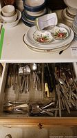 Top view of open kitchen drawer with assorted forks, spoons, knives, and other utensils arranged in dividers, along with decorative ceramic bowls and plates on countertop.