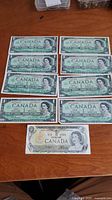 Nine Canadian one dollar bills displayed on a wooden surface: eight 1967 Centennial bills and one 1973 bill.