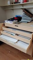 Wide view of flat file organizer with drawers open showing large sheets of art paper and assorted office supplies on top, with plastic bins and basket.