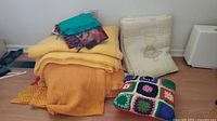 Stack of yellow blankets, colorful scarves on top, and cream quilt standing upright alongside a multicolored crochet pillow on the floor.