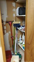 Photo of a basement closet showing red mop, blue broom with sponge, green and white handles, cleaning liquids on shelves.