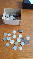 Coins spread on a wooden surface with a box containing additional coins in background, showcasing overall condition and variety of coins.