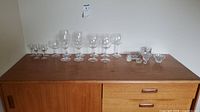 Wide shot of eleven clear crystal wine and aperitif glasses arranged in a line on a wooden surface, along with small crystal decorative pieces including a candle holder and a small dish.