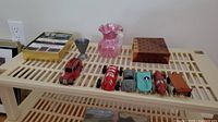 Wide view of all lot items displayed on a beige rack including six toy cars, a small pink pitcher, the address book, travel game box, and a small wooden box.