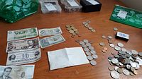 Top view of paper money and assorted coins arranged on wooden surface with some small boxes and green plastic bag in background.