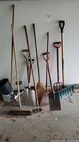 Full set of garden tools arranged side by side against wall including shovels, rakes, brooms, pitchfork and scoop