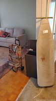 Ironing board with ripped beige cover, laundry hamper, and shopping cart in living room area.