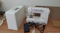Kenmore sewing machine alongside hard white plastic carrying case and black foot pedal with attached cord on light wood floor.