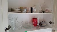 Cabinet shelf showing multiple kitchenware items including vases, bowls, glass plates, teapots, measuring cups and jugs.