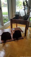 Photo of two wooden speaker stands and one wooden plant stand with some small potted plants inside the plant stand box, showing wood floor and window background.