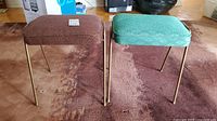 Two Mid-Century Modern stools with fabric seats and metal legs, one brown and one teal, positioned side-by-side on a carpeted floor.