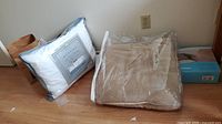 Photo showing a white microfiber down alternative pillow with a gray border still in packaging and a beige/tan blanket also sealed in plastic, placed on a floor against a wall.