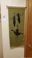 Full view of the hanging print on burlap showing two moose and kayaker in black silhouette on an olive background, hanging from a rod.