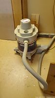Front and side views of the white and gray wet/dry shop vacuum with hose and wheeled base on a concrete floor.