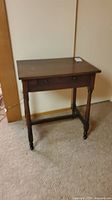 Front view of vintage wooden side table showing the drawer, turned legs, and casters.