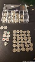Overview of multiple Canadian quarters stored in a clear compartment box and laid out on a black surface.