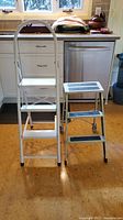 Front view of two folding step stools, white with handle and gray with anti-slip pads