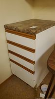 Four drawer white storage unit with wood trim handles and a faux marble top, located in a basement corner.