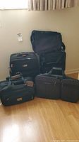 Photo of six black suitcases and bags assorted on wooden floor against wall, showing variety in size and handles.